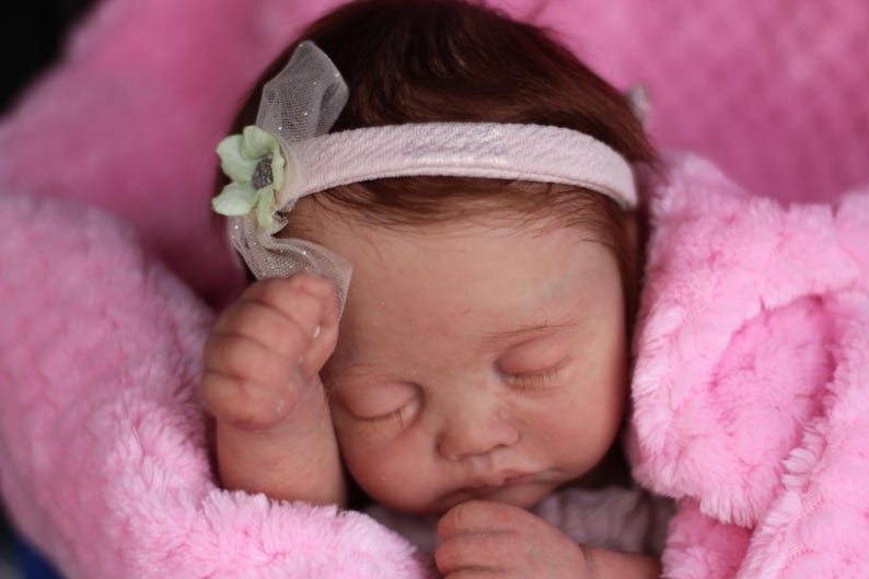 Puede incluir: Un mu&ntilde;eco beb&eacute; durmiendo con una diadema rosa y una flor verde. El mu&ntilde;eco lleva un vestido blanco y est&aacute; acostado en una manta rosa.