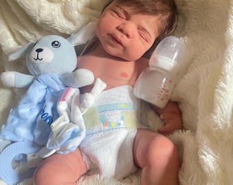 May include: A baby doll with brown hair, wearing a nappy, lies on a fluffy white blanket. A blue stuffed bunny and a baby bottle are next to the doll. The doll has a realistic appearance.