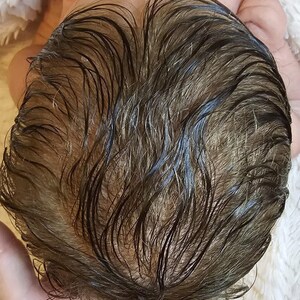 May include: Close-up of a baby's head with wet, dark brown hair. The hair is parted and shows the texture and color variations. The baby is lying on a white, fluffy surface.
