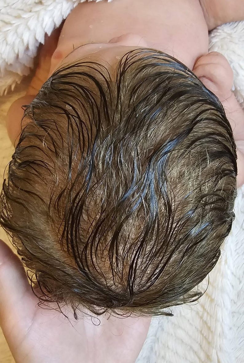 May include: Close-up of a baby's head with wet, dark brown hair. The hair is parted and shows the texture and color variations. The baby is lying on a white, fluffy surface.