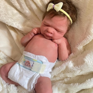 May include: A lifelike baby doll with brown hair and a yellow headband with a bow. The doll is wearing a diaper with a colorful design and is lying on a soft, white blanket. The doll's skin has a realistic tone.