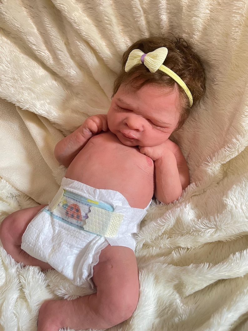 May include: A lifelike baby doll with brown hair and a yellow headband with a bow. The doll is wearing a diaper with a colorful design and is lying on a soft, white blanket. The doll's skin has a realistic tone.