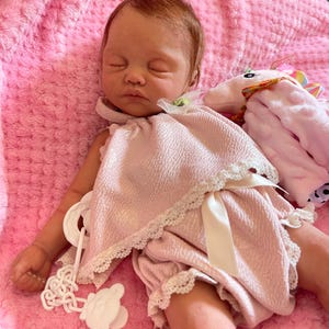 Puede incluir: Un beb&eacute; reborn durmiendo que lleva un vestido rosa y bombachos a juego con ribete de encaje blanco. El beb&eacute; est&aacute; tumbado en una manta rosa peluda con un animal de peluche de unicornio.