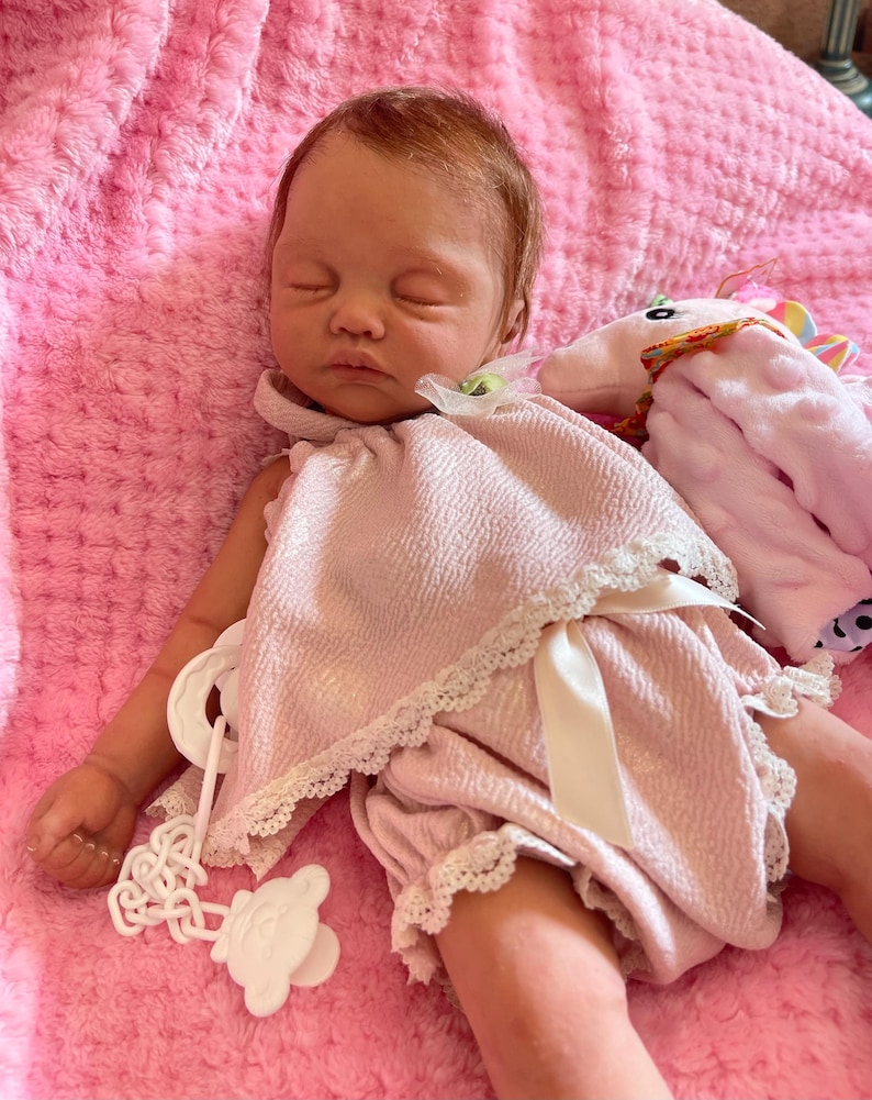 Puede incluir: Un beb&eacute; reborn durmiendo que lleva un vestido rosa y bombachos a juego con ribete de encaje blanco. El beb&eacute; est&aacute; tumbado en una manta rosa peluda con un animal de peluche de unicornio.