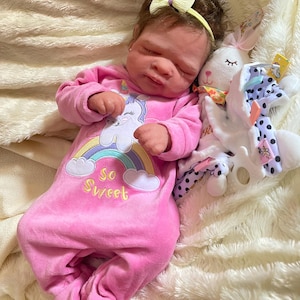 May include: A baby doll wearing a pink onesie with a unicorn and rainbow design, the text "So Sweet", and a yellow bow headband. A white stuffed bunny with a patterned scarf and a polka dot blanket are next to the doll.
