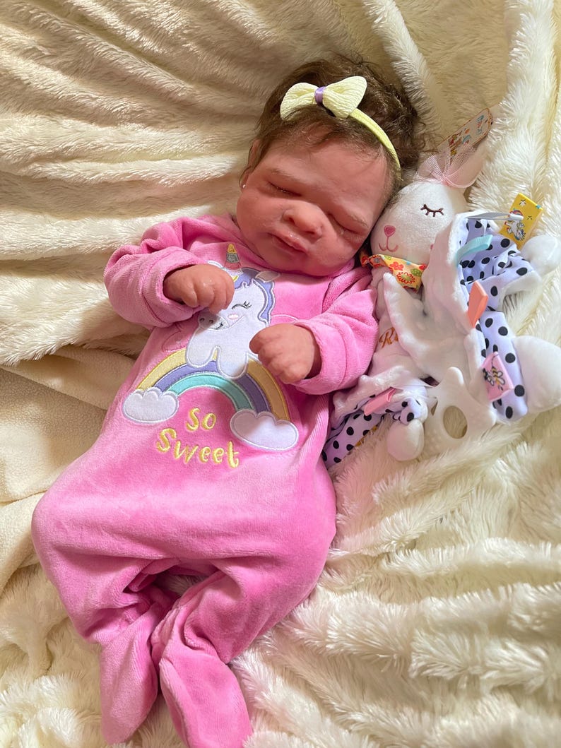 May include: A baby doll wearing a pink onesie with a unicorn and rainbow design, the text "So Sweet", and a yellow bow headband. A white stuffed bunny with a patterned scarf and a polka dot blanket are next to the doll.