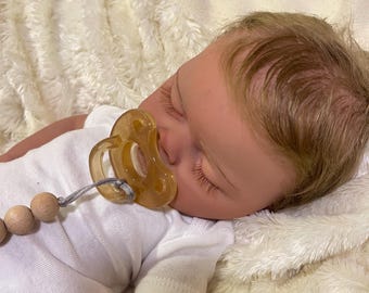 Puede incluir: Un muñeco de bebé durmiendo con cabello rubio, vistiendo un mameluco blanco y un chupete. El muñeco está acostado sobre una manta blanca y esponjosa.