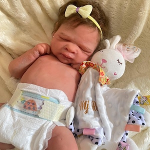 May include: A realistic baby doll with brown hair and a yellow bow headband, wearing a diaper. The doll is lying next to a white bunny-shaped security blanket with a pink bow and polka dot accents.