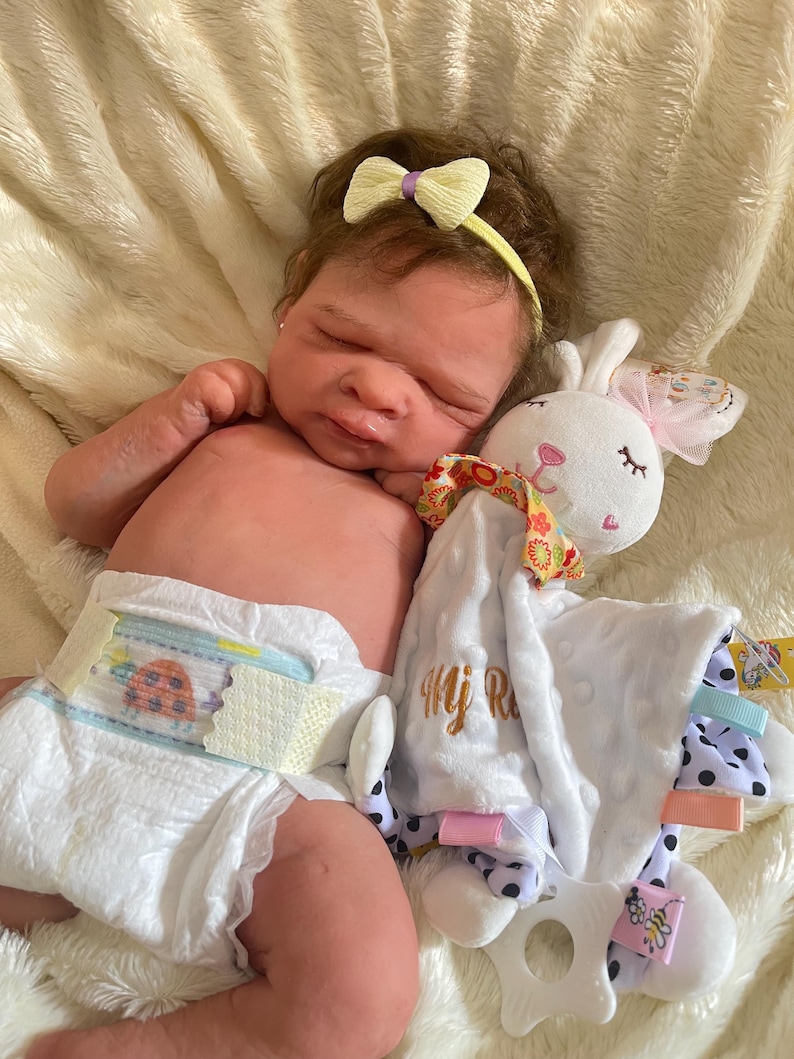 May include: A realistic baby doll with brown hair and a yellow bow headband, wearing a diaper. The doll is lying next to a white bunny-shaped security blanket with a pink bow and polka dot accents.