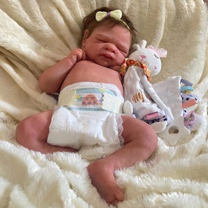 May include: A realistic baby doll with a light skin tone, wearing a diaper with a ladybug design. The doll has a yellow bow headband and is lying on a fluffy white blanket. A stuffed animal is next to the doll.