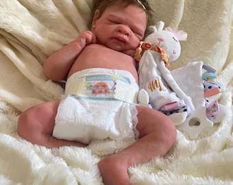 Puede incluir: Una muñeca bebé realista con piel clara, que lleva un pañal con un diseño de mariquita. La muñeca tiene una diadema con lazo amarillo y está acostada sobre una manta blanca y esponjosa. Un peluche está al lado de la muñeca.