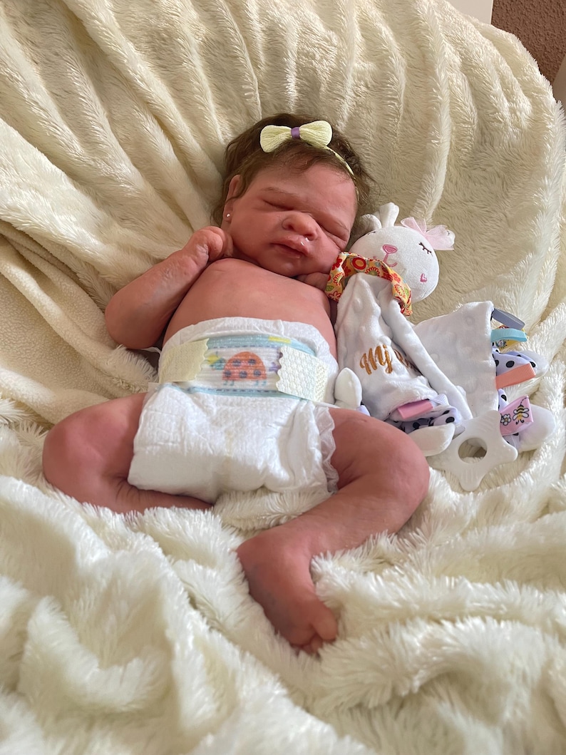 May include: A realistic baby doll with a light skin tone, wearing a diaper with a ladybug design. The doll has a yellow bow headband and is lying on a fluffy white blanket. A stuffed animal is next to the doll.