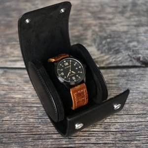 Könnte beinhalten: Eine schwarze Lederuhrenrolle mit einer Uhr mit braunem Lederarmband im Inneren. Das Zifferblatt der Uhr ist schwarz mit weißen Markierungen und dem aufgedruckten Text "OCCHISTA AUTOMATIC".