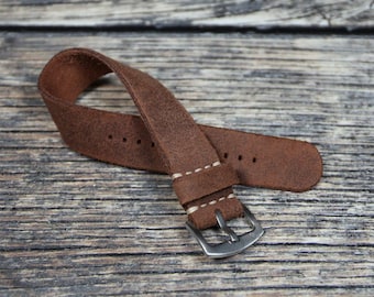 May include: A brown leather watch strap with white stitching and a silver buckle. The strap is folded in half and lying on a wooden surface.