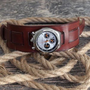Peut inclure: Une montre avec un bracelet manchette en cuir marron et un cadran argenté. Le cadran de la montre présente des détails noirs et orange. Le bracelet est posé sur une corde.