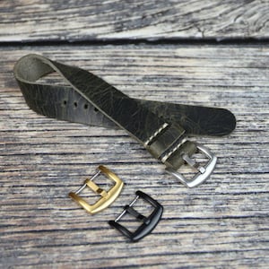 Puede incluir: Una correa de reloj de cuero verde oscuro con costuras blancas y una hebilla plateada. Se muestran dos hebillas adicionales, una dorada y una negra.