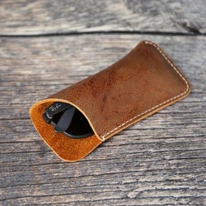 May include: A brown leather sunglasses case with white stitching. The case is closed and a pair of sunglasses is visible inside.