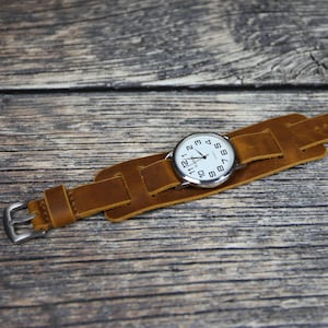 Puede incluir: Una correa de reloj de cuero marrón con una esfera plateada. La esfera del reloj tiene números y manecillas negros.