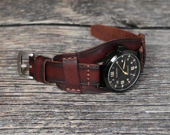 May include: A brown leather watch strap with a black watch face. The watch face has white numbers and a gold second hand. The strap is designed to wrap around the wrist.