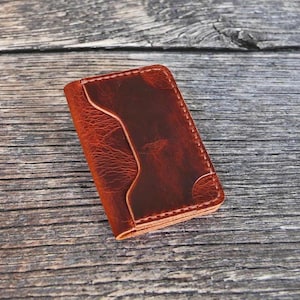 Può includere: Un piccolo portafoglio in pelle fatto a mano, nei toni del marrone. Il portafoglio presenta un design unico con una tasca curva e cuciture a vista. La pelle ha un aspetto invecchiato, che aggiunge carattere all'accessorio.