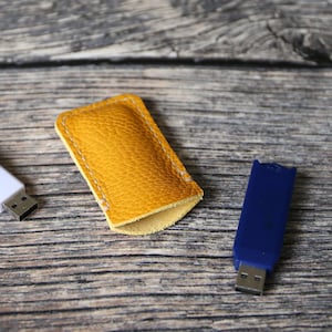 May include: A yellow leather case with white stitching for a USB flash drive. A blue USB flash drive is also pictured on a wooden surface.