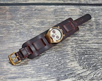 May include: A gold-toned wristwatch with a cream-colored face and red sub-dials is set on a wide, dark brown leather cuff. The cuff features a gold-colored buckle and is designed to wrap around the wrist. The watch and cuff are displayed on a wooden surface.