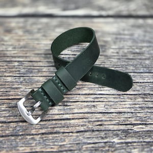 Op de afbeelding: Een donker bosgroene lederen horlogeband met een matte afwerking, twee bijpassende lederen lussen en een gesp van geborsteld zilverkleurig metaal. De band heeft zichtbare stiksels langs de randen en meerdere verstelgaatjes aan het uiteinde.