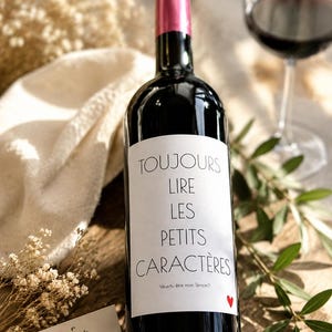 Peut inclure: Une bouteille de vin en verre foncé avec une capsule rose et une étiquette blanche portant le texte noir TOUJOURS LIRE LES PETITS CARACTÈRES, la mention Veux-tu être mon témoin ? et un petit cœur rouge dans le coin inférieur droit.