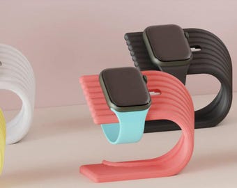 Puede incluir: Cuatro soportes de diferentes colores para Apple Watch. Cada soporte está hecho de un material texturizado, flexible y tiene una forma curva. Los soportes son de color blanco, amarillo, negro y rosa. Los Apple Watch son de color negro con correas de diferentes colores: blanco, azul, negro y verde.