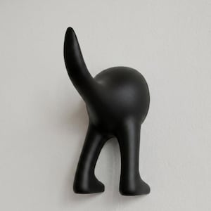 Könnte beinhalten: Ein schwarzer Wandhaken in Form eines Hundes. Der Haken hat einen abgerundeten Körper, zwei Beine und einen nach oben zeigenden Schwanz. Er ist an einer weißen Wand befestigt. Ein dekoratives und funktionales Objekt.