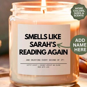May include: A white candle with a label that reads "Smells like Sarah's Reading Again...and enjoying every second of it!"  Gift it Give it | Coconut Apricot Wax Blend | Made with Love.  The label also has a space to add a name.  The candle is on a wooden surface.