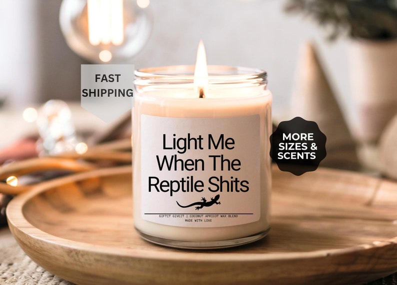 K&ouml;nnte beinhalten: Eine wei&szlig;e Kerze mit einem schwarzen Etikett, auf dem "Light Me When The Reptile Shits" steht. Das Etikett zeigt auch eine kleine schwarze Eidechsengrafik. Die Kerze steht auf einem Holztablett.