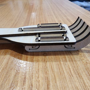 K&ouml;nnte beinhalten: Ein h&ouml;lzerner Spielzeugschlitten mit einer wei&szlig;en Basis und braunen Akzenten. Der Schlitten besteht aus mehreren Holzschichten und hat eine geschwungene Form.