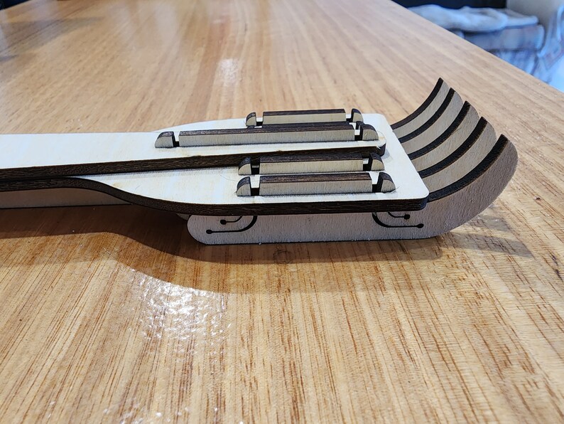 K&ouml;nnte beinhalten: Ein h&ouml;lzerner Spielzeugschlitten mit einer wei&szlig;en Basis und braunen Akzenten. Der Schlitten besteht aus mehreren Holzschichten und hat eine geschwungene Form.