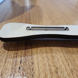 K&ouml;nnte beinhalten: Nahaufnahme eines Gitarren-Stegs aus Holz mit dunkelbrauner Oberfl&auml;che. Der Steg besteht aus zwei Holzst&uuml;cken mit einer Nut in der Mitte f&uuml;r die Saiten.