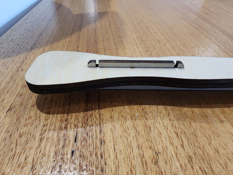 K&ouml;nnte beinhalten: Nahaufnahme eines Gitarren-Stegs aus Holz mit dunkelbrauner Oberfl&auml;che. Der Steg besteht aus zwei Holzst&uuml;cken mit einer Nut in der Mitte f&uuml;r die Saiten.