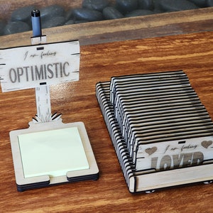 May include: Two wooden note holders with engraved messages. The first holder has a sign that reads "I am feeling OPTIMISTIC" and a notepad. The second holder has a sign that reads "I am feeling LOVED" with two hearts.