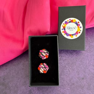 Könnte beinhalten: Ein Paar abstrakte geometrische Ohrringe in einer schwarzen Schachtel. Die Ohrringe weisen ein Muster aus rosa, roten und weißen Formen auf. Die Schachtel ist geöffnet und zeigt die Ohrringe vor einem schwarzen Hintergrund. Eine zweite geschlossene Schachtel mit einem farbenfrohen Logo ist sichtbar.