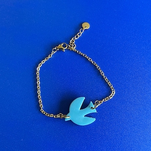 Può includere: Un braccialetto a catena color oro con un ciondolo a forma di uccello azzurro. Il braccialetto ha una catena delicata e un piccolo ciondolo rotondo all'estremità. Il ciondolo a forma di uccello è azzurro.