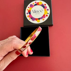 Peut inclure: Un bracelet jonc doré avec un motif tacheté blanc et rose. Le bracelet est présenté dans un coffret noir avec le nom de la marque "Alinoé Creations" sur le couvercle. Le fond est rouge uni.