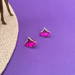 Peut inclure: Deux boucles d'oreilles à clous florales roses et blanches sur fond violet. Les boucles d'oreilles ont une forme de ventilateur avec un motif floral rose et des accents noirs et blancs. Les boucles d'oreilles sont petites et délicates.