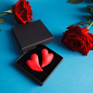Puede incluir: Dos caramelos rojos en forma de corazón en una caja de regalo negra con una rosa roja sobre un fondo azul.