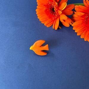Peut inclure: Une broche en forme d'oiseau orange vif avec un bec jaune, posée sur un fond bleu profond, avec deux gerberas orange vibrantes. La broche a un design simple et stylisé. Les fleurs ont de nombreux pétales et un centre sombre.