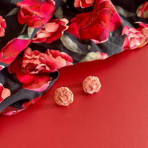 Peut inclure: Deux petites boucles d'oreilles roses en forme de rose sont posées sur une surface rouge à côté d'un tissu noir avec un grand motif floral rouge et rose. Le tissu est plié, montrant le design complexe des fleurs.