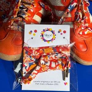 Peut inclure: Une paire de baskets orange à bandes blanches avec des lacets à motifs floraux, présentée avec un lot de lacets en tissu de 135 cm sur une carte blanche avec le logo Alinoé et le texte Lacets originaux en 135 cm Fait main à Redon (35).
