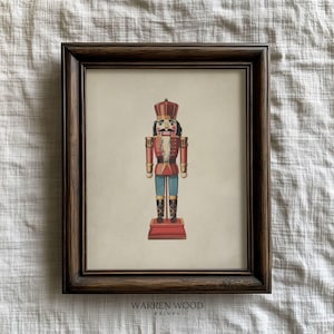 May include: Framed print of a traditional nutcracker soldier in red, blue, and black. The print is set against a cream background and framed in a dark brown wooden frame. The text "WARREN WOOD PRINTS" is at the bottom.