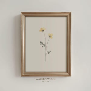 May include: A framed print featuring a minimalist botanical design. Two delicate yellow flowers with green leaves are centered on a beige background. The print is in a gold-colored frame. The text "WARREN WOOD PRINTS" is at the bottom.