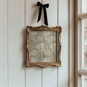 May include: A gold-toned ornate picture frame with a floral pattern inside. The frame is hanging on a white wall with a black bow at the top.