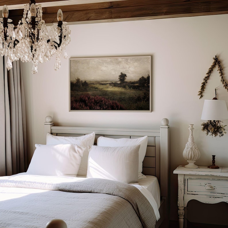 May include: A bedroom scene featuring a bed with white bedding and pillows, a framed landscape painting, and a crystal chandelier. A white nightstand with a lamp and decorative elements sits beside the bed. The room has a neutral color palette.