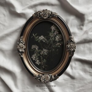 May include: An oval framed print featuring a floral botanical design. The frame is black and gold with ornate floral accents. The print has a dark background with white flowers and green leaves. The text "WARREN WOOD PRINTS" is at the bottom.